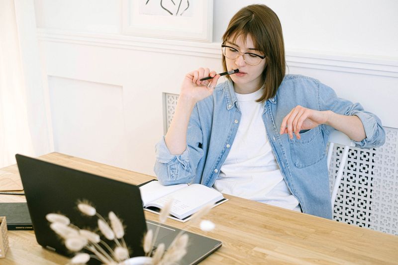 A woman holding a pen thinking in front of a laptop that's on a table